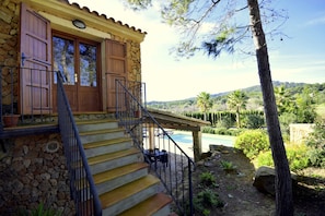 A stone house with a wooden staircase leading to a balcony overlooking a pool and lush greenery. - 44246 Finca Sa Cova, Spain (Petra)