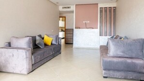 A modern living room with two grey sofas, a pink accent wall, and a visible kitchen area in the background.