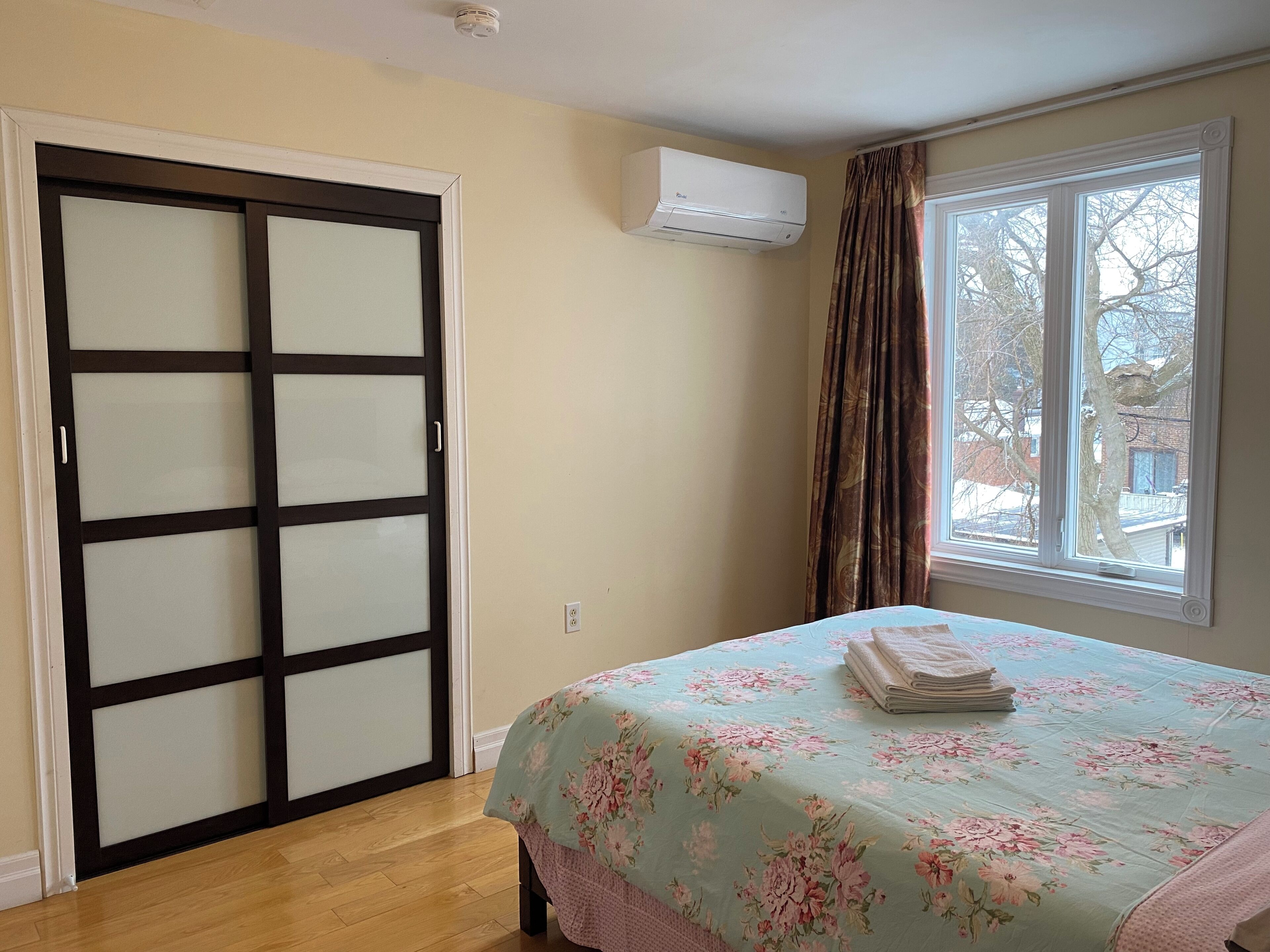 Un dormitorio con cama, ventana con cortinas, aire acondicionado de pared y puerta corrediza.