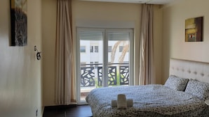A hotel room with a bed, a small table, a painting on the wall, and a balcony with a view of buildings and palm trees.