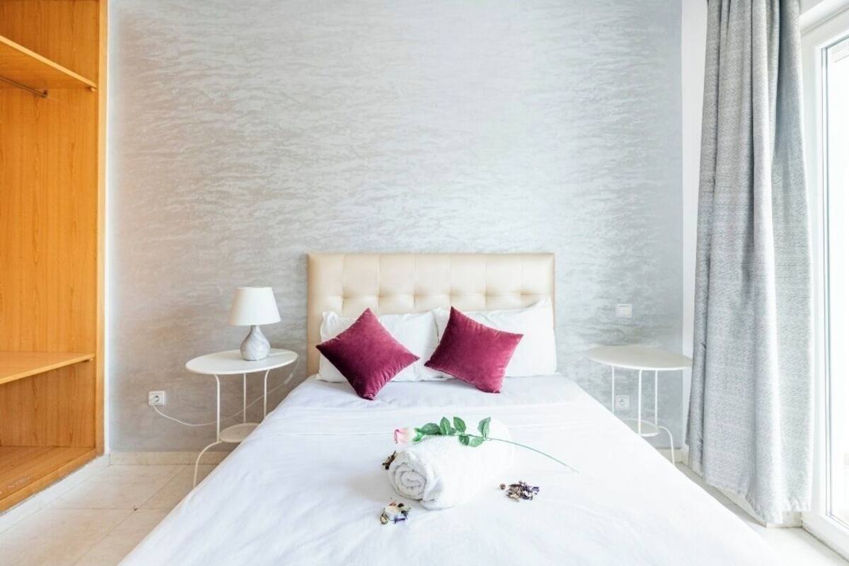 Ein ordentlich bezogenes Bett mit weißer Daunendecke, zwei violetten Kissen und einem eingerollten Handtuch mit einem grünen Blatt. Neben dem Bett steht ein kleiner weißer Tisch mit einer Lampe.