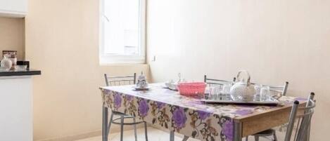 Un comedor con una mesa preparada para cuatro, una ventana con persianas y una zona de cocina a la izquierda.