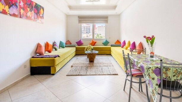 A bright living room with a yellow sofa, colorful cushions, a coffee table, and a dining table with chairs. - Appartement 15 ensoleillé à 5 min de la plage El Jadida (El Jadida)