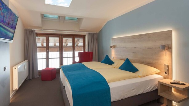A hotel room with a large bed, a red chair, a TV, a skylight, and a window with curtains. - Dachappartement (Berchtesgaden)