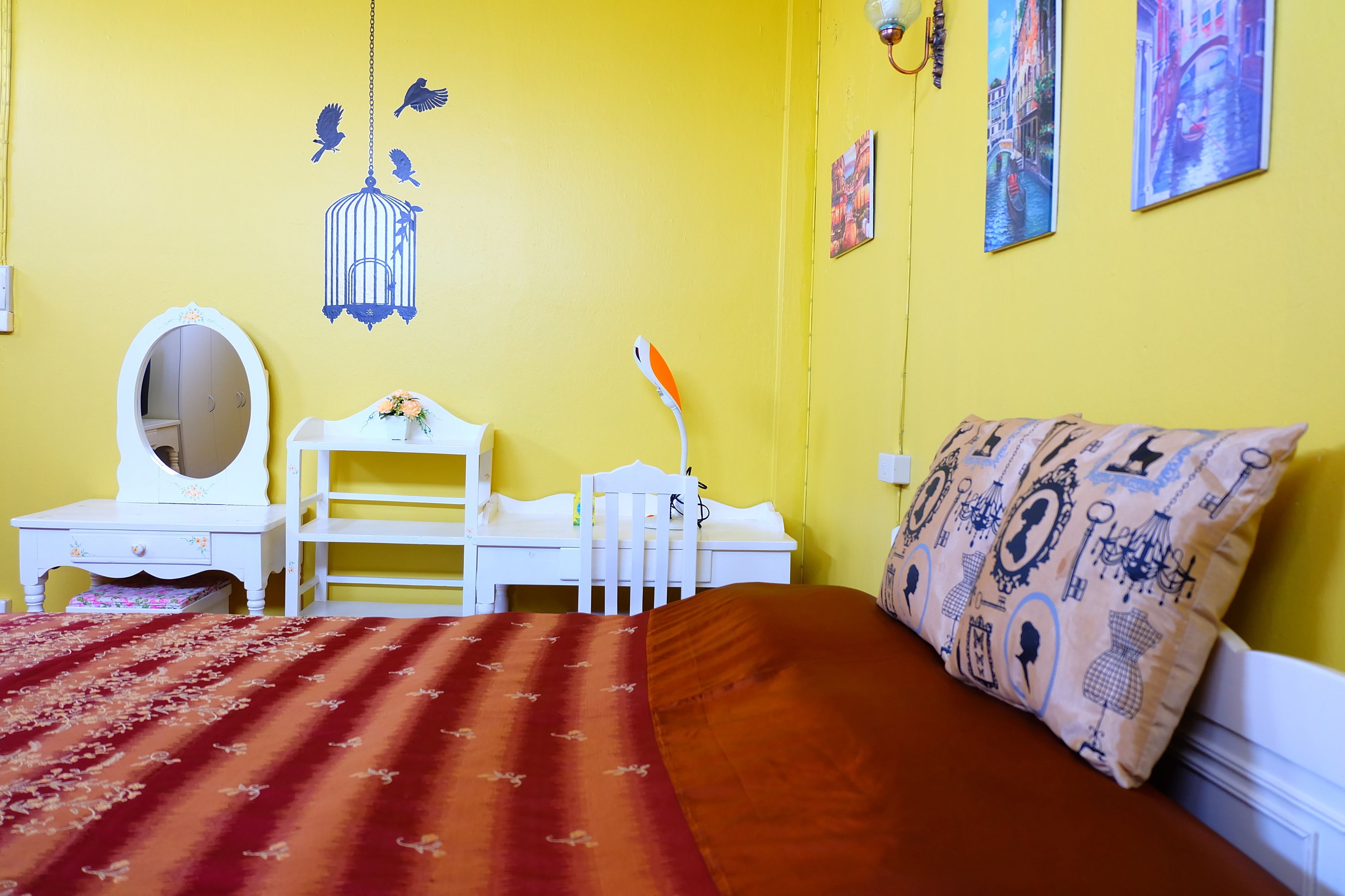 Una habitación con una pared amarilla, una cama con un cojín estampado, un aparador con espejo y una jaula colgante.