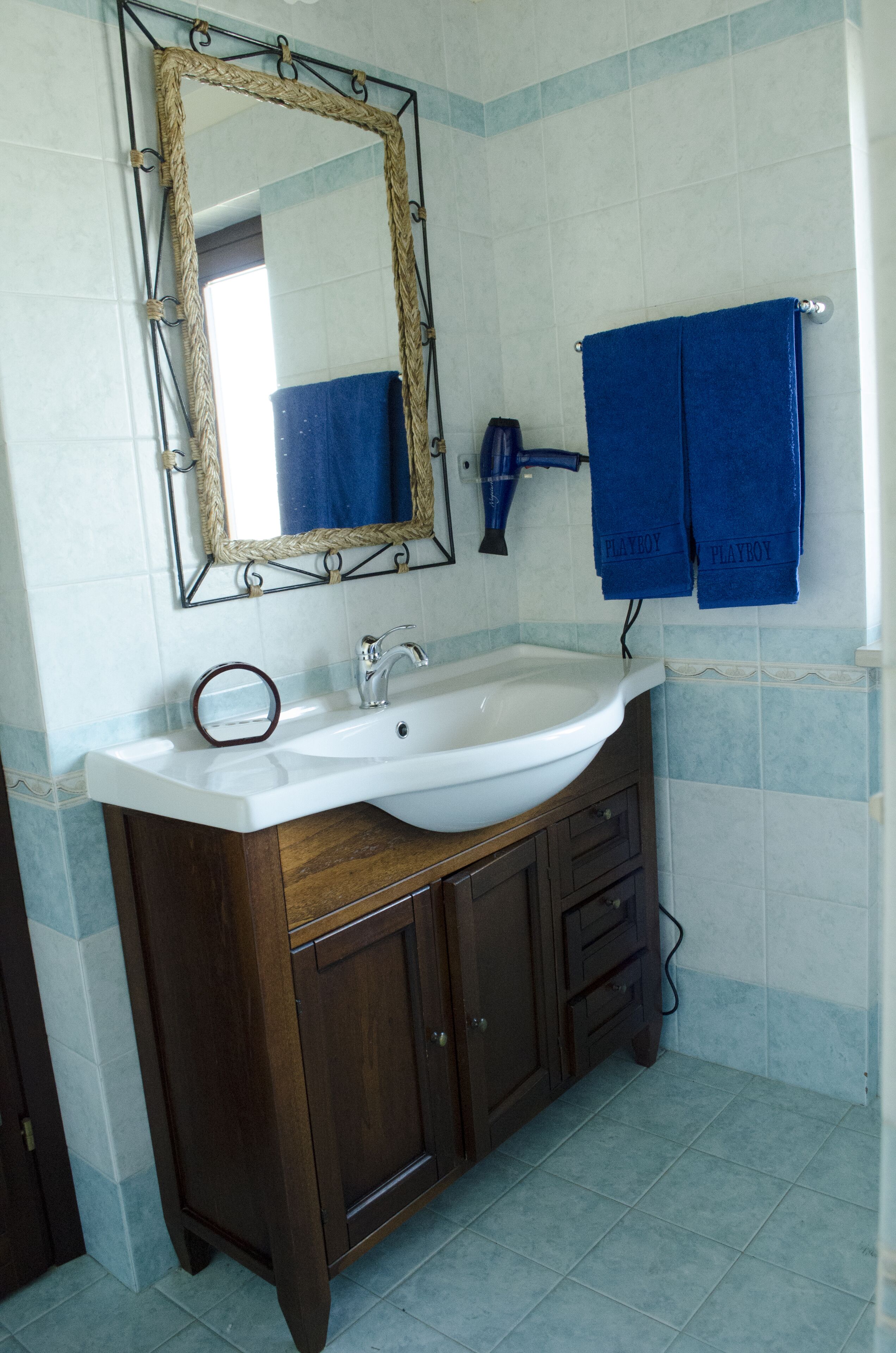 Un baño con un lavamanos blanco, tocador de madera, un espejo con marco decorativo y dos toallas azules colgadas en un perchero.