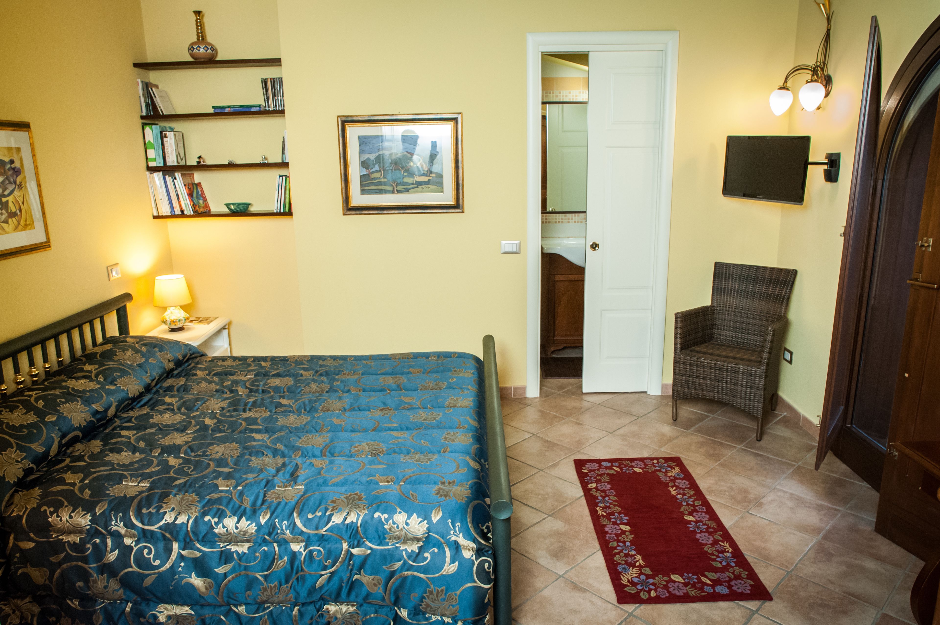 A bedroom with a bed, a chair, a bookshelf, a framed picture, and a door leading to another room.