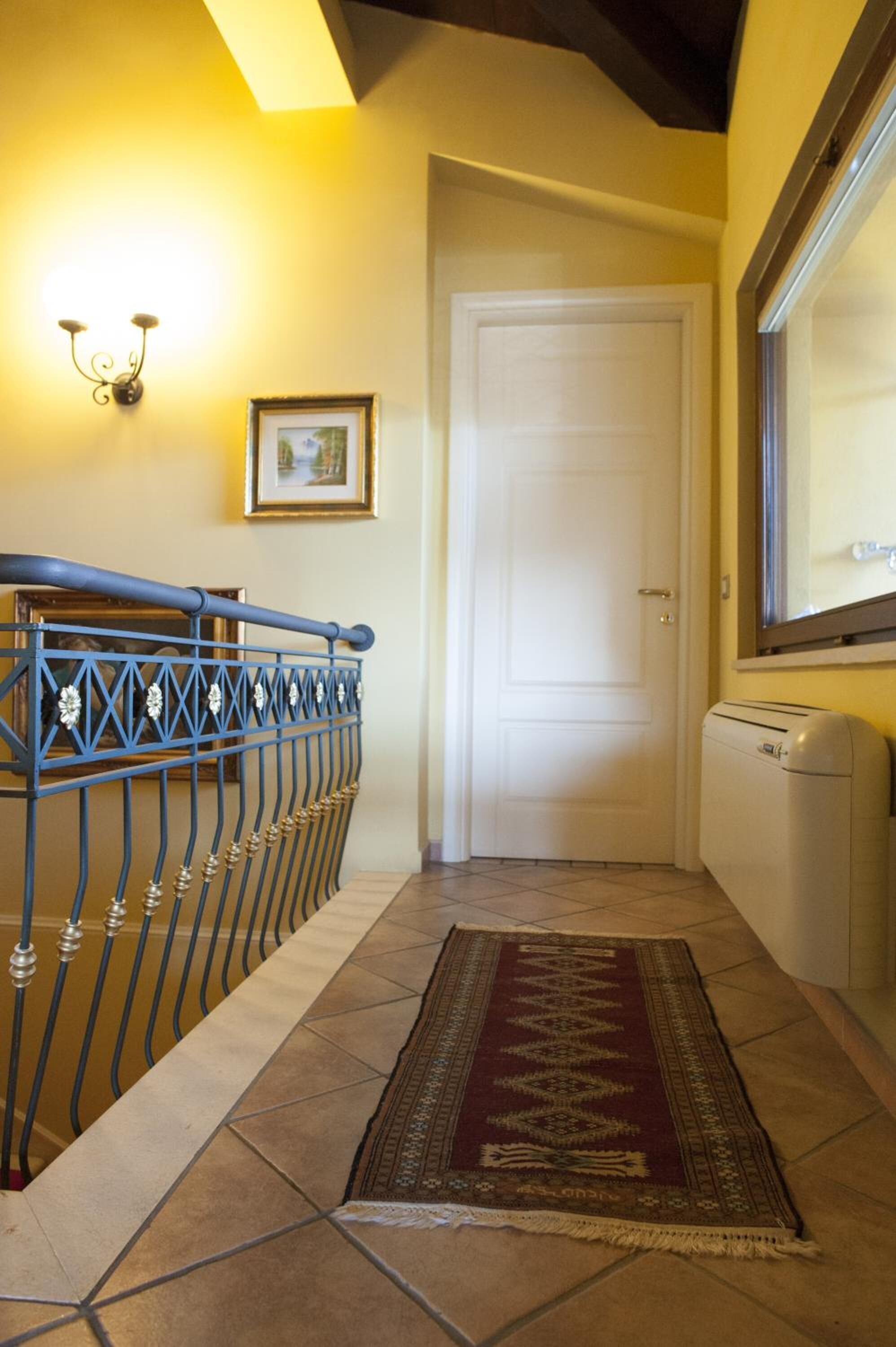 A hallway with a blue metal railing, a white door, a framed picture on the wall, and a patterned rug on the floor.