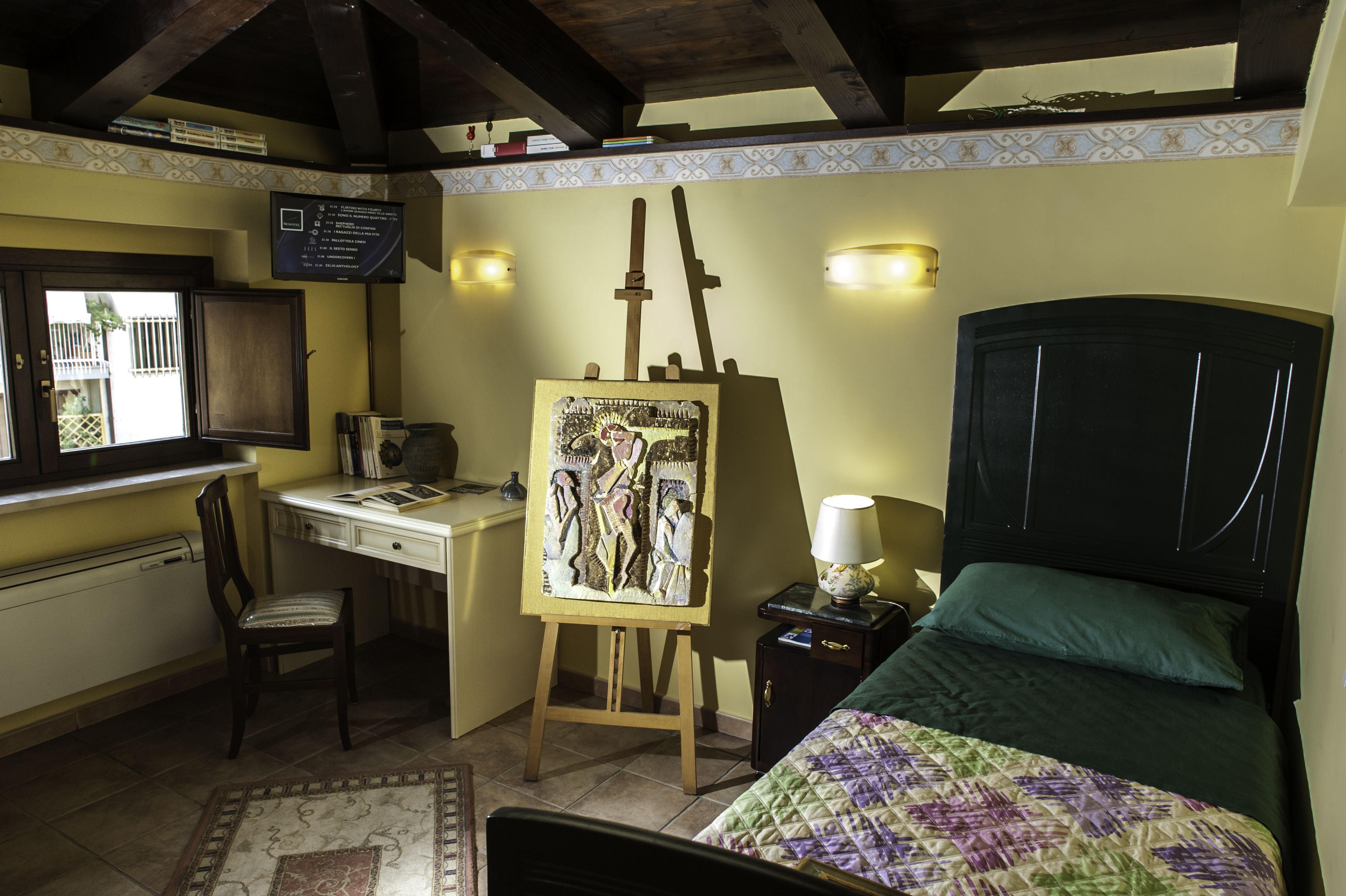 Ein Schlafzimmer mit einem Bett, einem Schreibtisch, einem Stuhl, einer Staffelei mit einem Gemälde und einem Fernseher an der Wand.