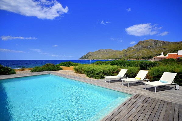 A pool with lounge chairs, a wooden deck, and a view of the ocean and mountains.