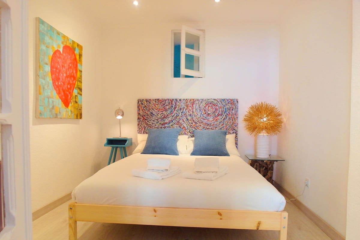 A bedroom with a wooden bed, a colorful abstract painting, a small window, and a decorative golden sphere on a side table.