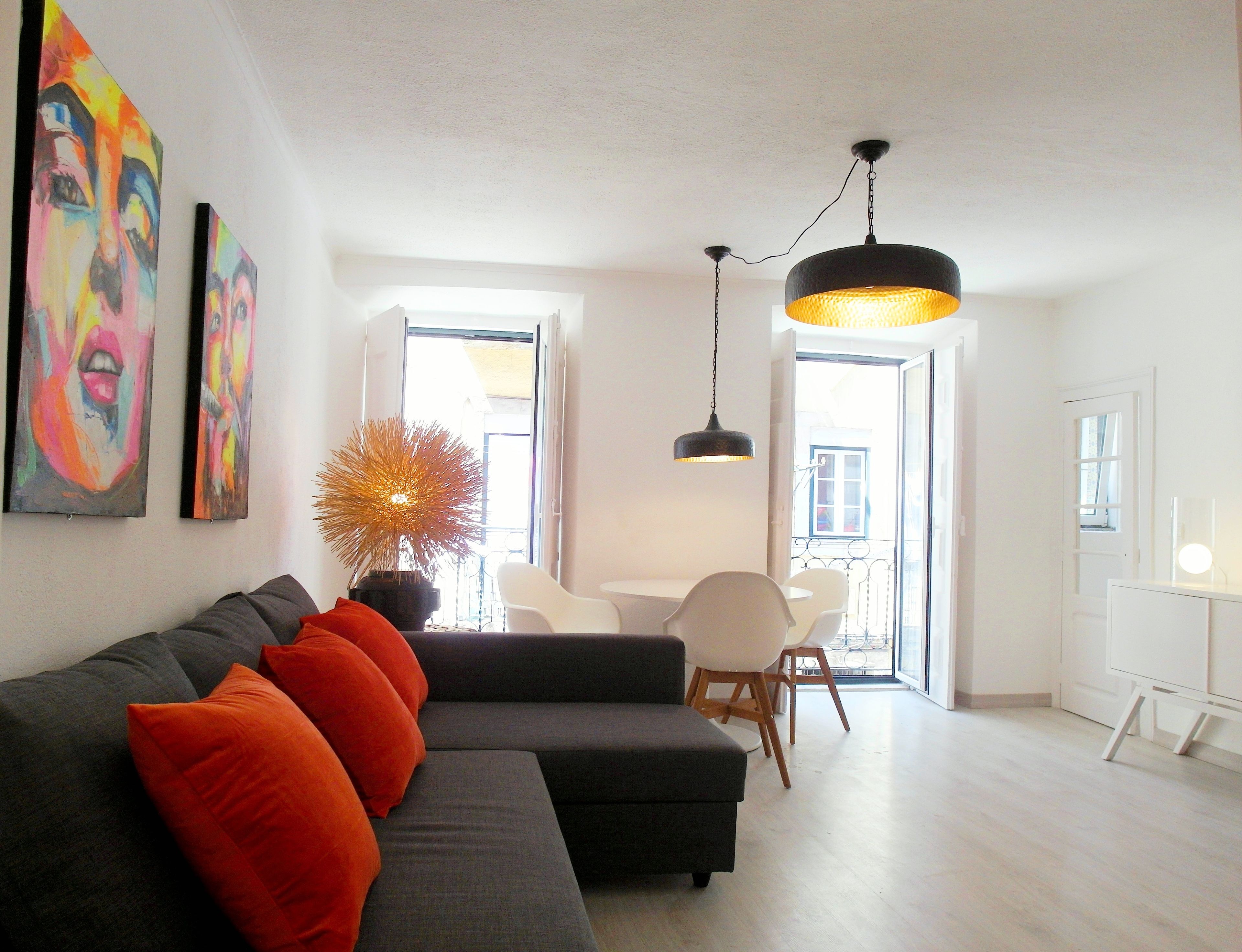 Ein modernes Wohnzimmer mit einem grauen Sofa, orangefarbenen Kissen, einer Topfpflanze und einem Essbereich mit weißen Stühlen und einem Tisch.