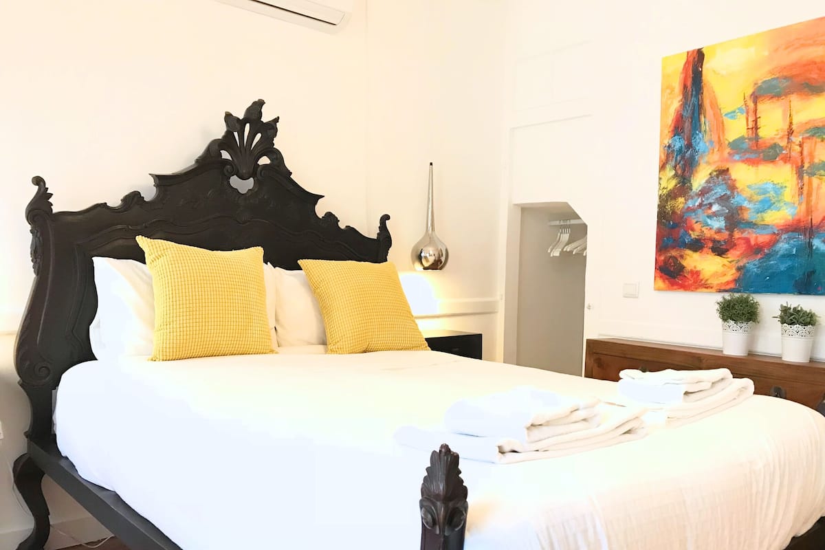 A bed with a black headboard, white bedding, and two yellow pillows. There is a painting on the wall and two potted plants on the side table.