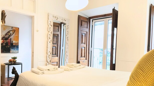 A neatly made bed with a white comforter and a yellow pillow, a wooden door with a mirror, and a painting on the wall. - SantoSweethome 2 (Lisbon)