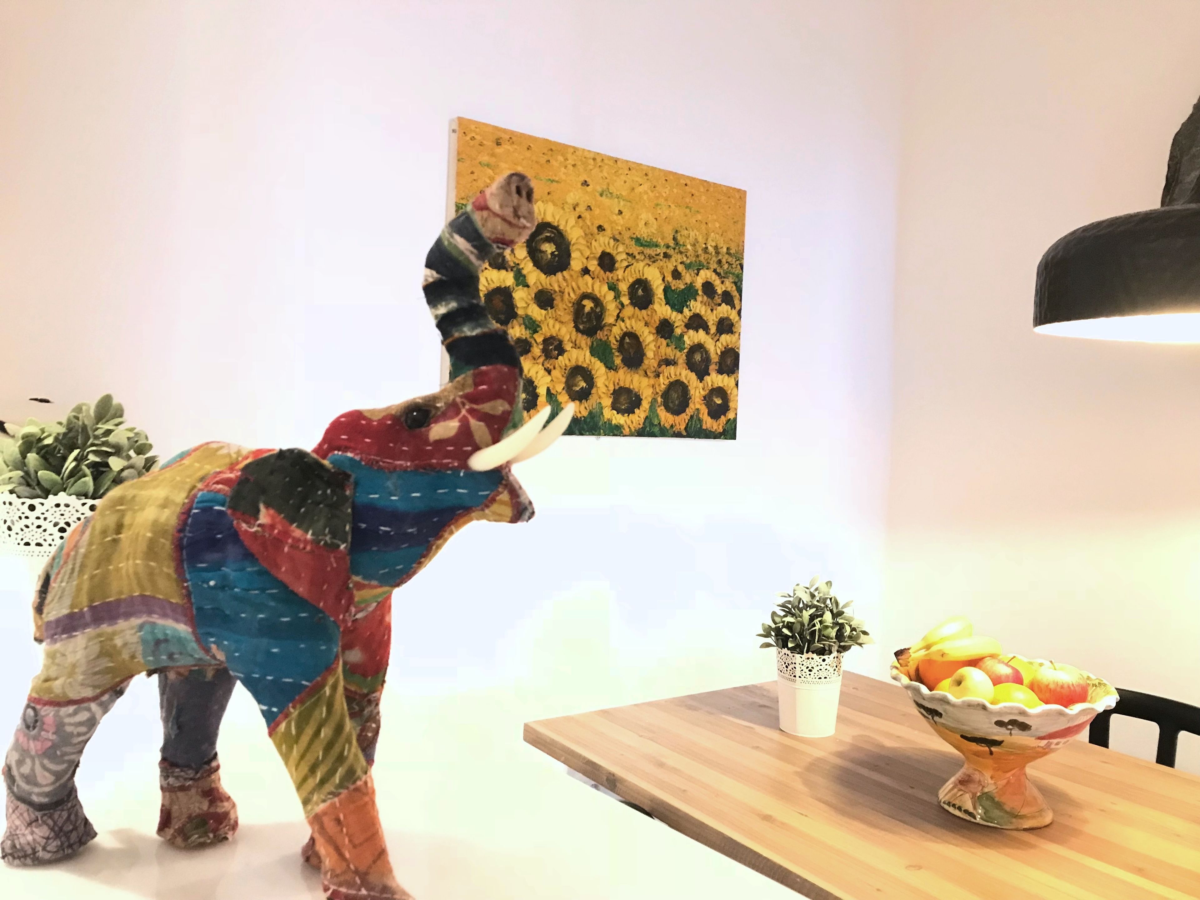 A colorful elephant sculpture in a dining area with a sunflower painting on the wall, a small potted plant, and a fruit bowl on the table.