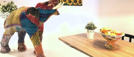 A colorful elephant sculpture in a dining area with a sunflower painting on the wall, a small potted plant, and a fruit bowl on the table.