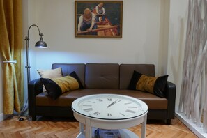 A living room with a brown sofa, a round table with a clock, a painting on the wall, and a floor lamp.