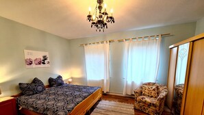 Un dormitorio con cama, una silla, un espejo, una araña y una ventana con cortinas.