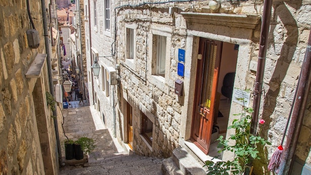 Une rue étroite bordée d’immeubles en pierre, une porte bien visible avec une enseigne, et un petit jardin agrémenté d’un rosier.
