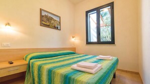 Um quarto com cama, mesa de cabeceira, gaveta, luminária na parede, janela e quadro pendurado na parede.