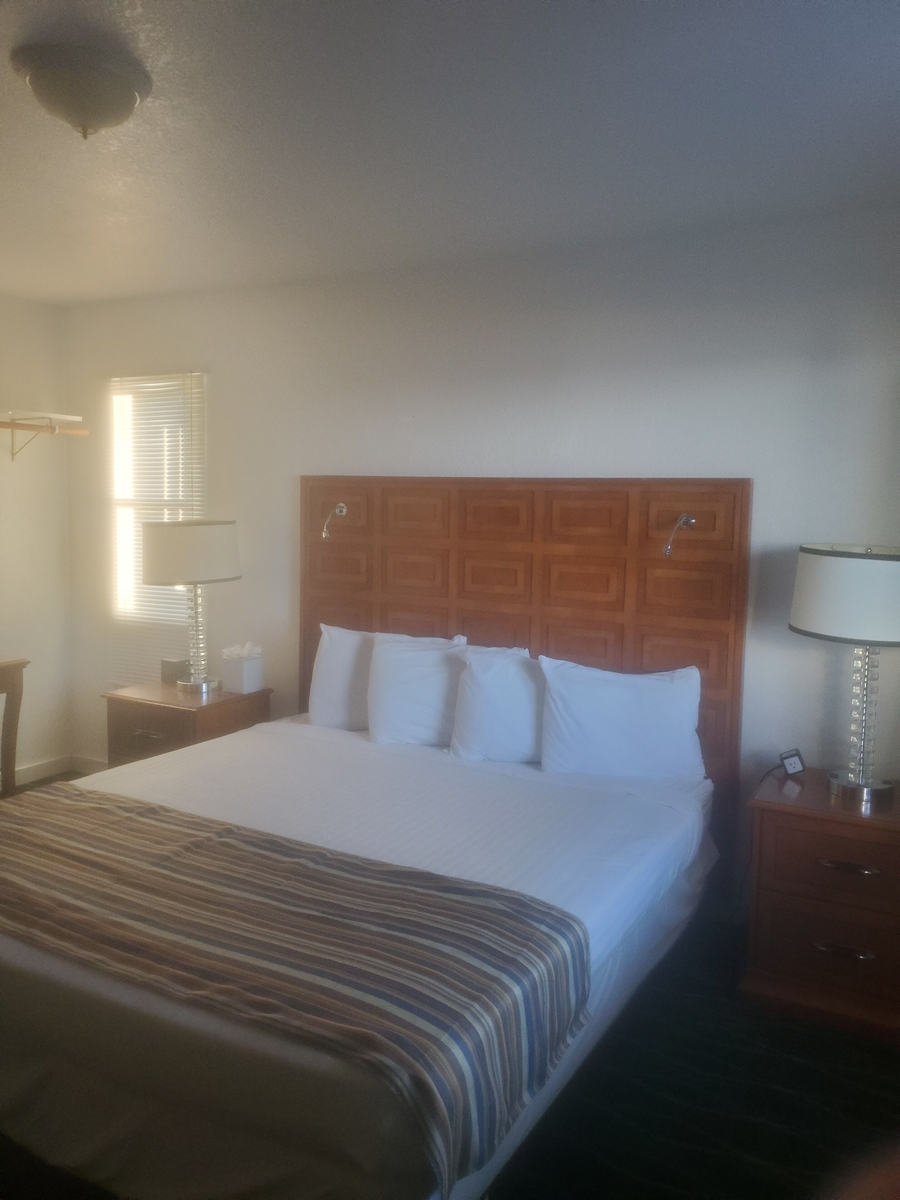 Una habitación de hotel con una cama grande, cabecero de madera, mesitas de noche con lámparas y una ventana con persianas.