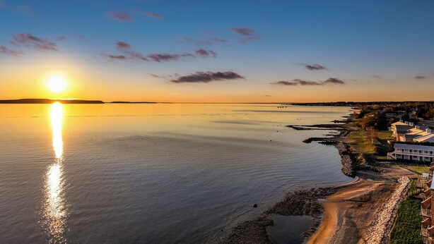 A coastal view with a sunset over the water, a rocky shoreline, and buildings along the coast. - Bayview Lakefront Motel (St Ignace)