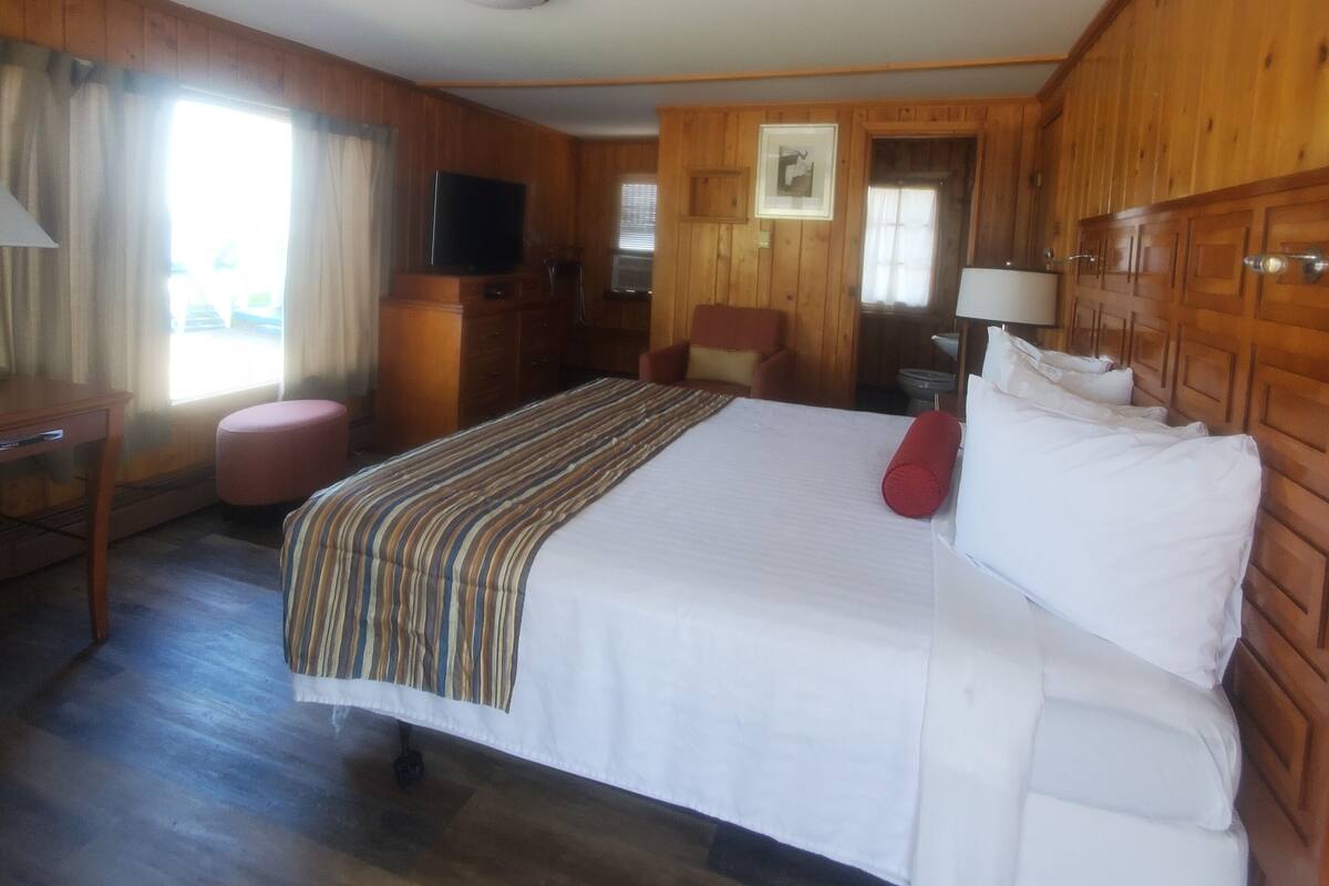 Quarto de hotel com uma cama grande, paredes de madeira, televisão, escrivaninha e janela com cortinas.