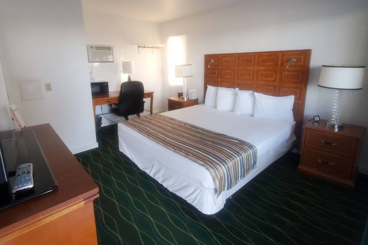 Quarto de hotel com uma cama grande, uma escrivaninha com cadeira, uma mesinha de cabeceira com abajur e um painel de controle na escrivaninha.