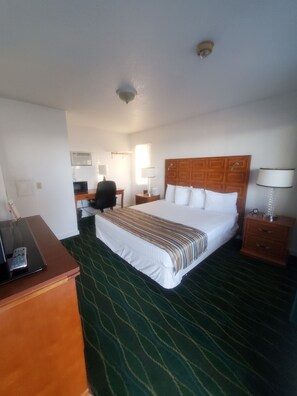 A hotel room with a large bed, a desk with a chair, a bedside table with a lamp, and a control panel on the desk. - Bayview Lakefront Motel (St Ignace)