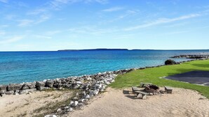 A coastal view with a rocky shoreline, a grassy area with benches, and a clear blue sky. - Bayview Lakefront Motel (St Ignace)
