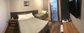 A hotel room with a large bed, a smaller bed, a bedside table with a lamp, a TV mounted on the wall, and a bathroom visible through an open door. - Hotel Blumenhof Balneário Camboriú (Balneário Camboriú)