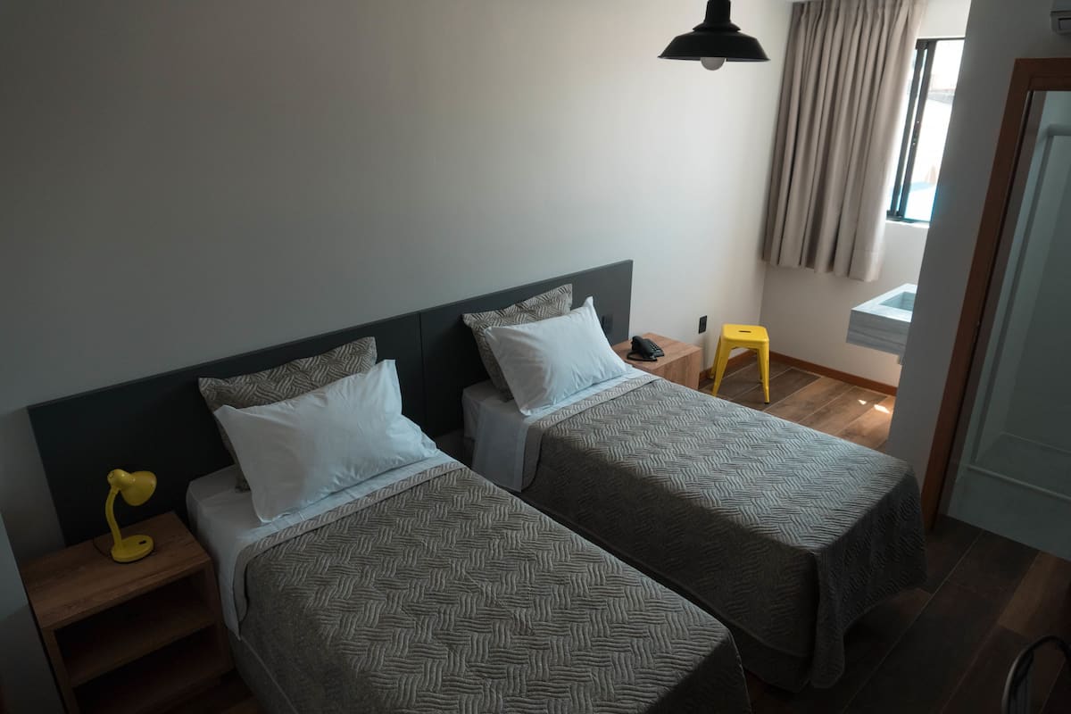Ein Hotelzimmer mit zwei Betten, einem Nachttisch mit Lampe, einem kleinen Hocker und einem Fenster mit Vorhängen.