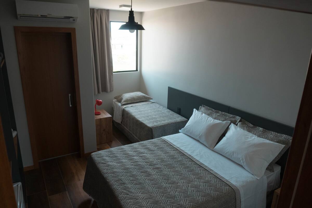 Ein Schlafzimmer mit zwei Betten, einem Schreibtisch mit Lampe, einem Fenster mit Vorhängen und einer Klimaanlage.