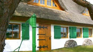 Une maison traditionnelle à toit de chaume, avec une porte en bois et des volets verts, une structure métallique verte à l’avant et des murs en pierre.