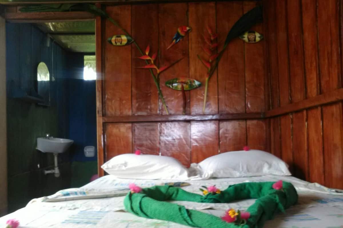 A wooden bed with white pillows and a green blanket, a wooden headboard with a floral design, and a sink in the adjacent room.