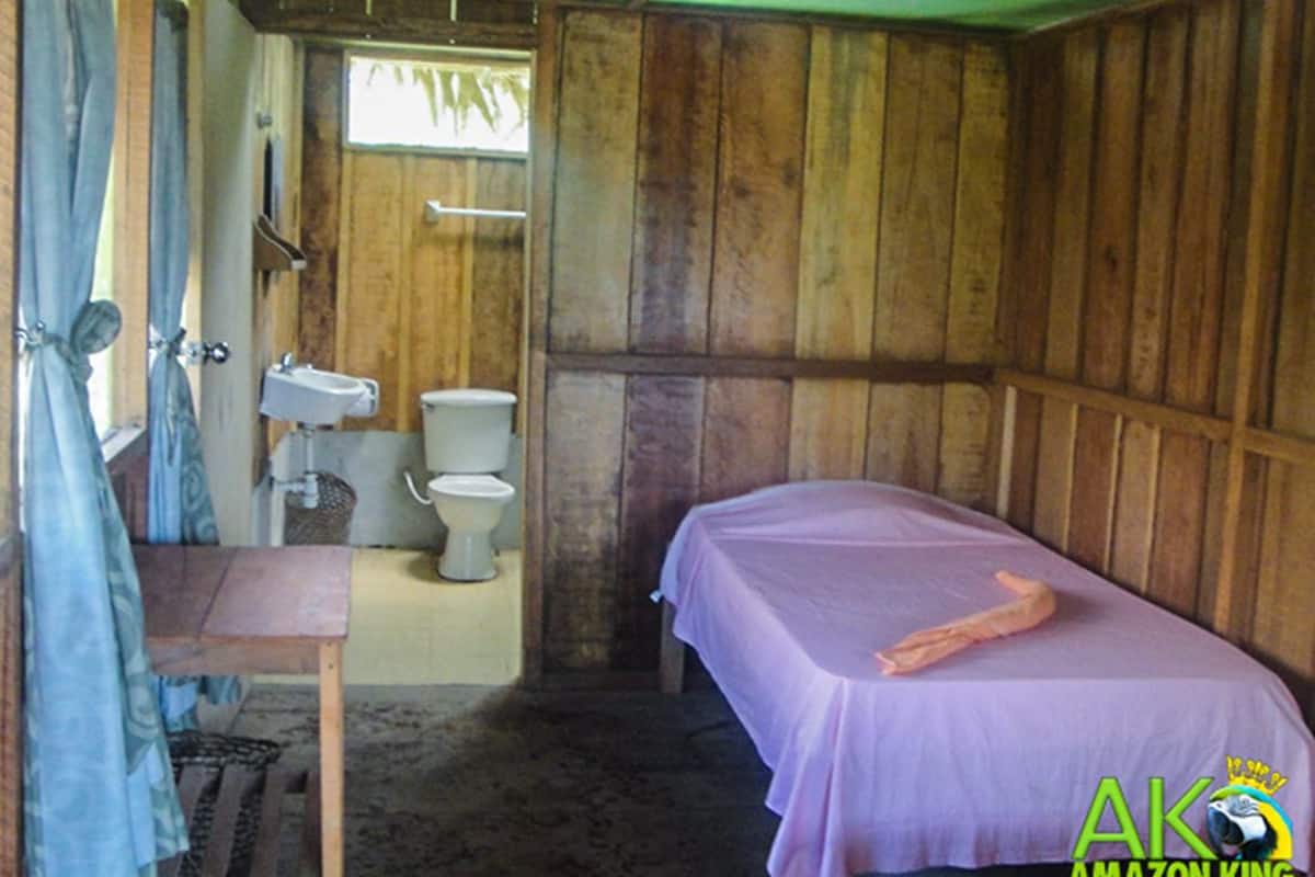 A room with wooden walls, a bed with a pink bedspread, a small table, a sink, and a toilet.