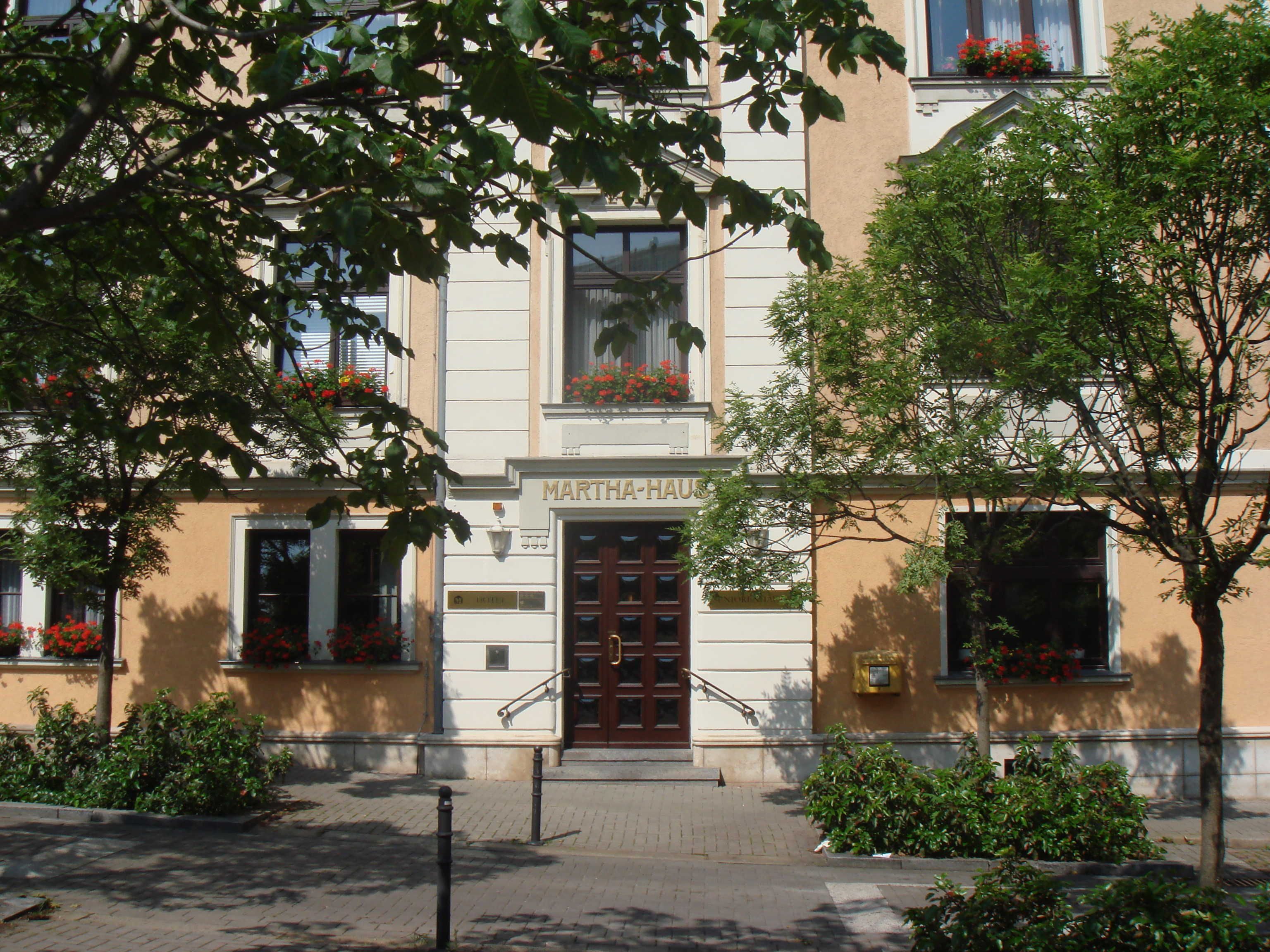 Ein Gebäude mit einem Schild, auf dem 'MARTHA-HAUF' steht, am Eingang, mit traditioneller Architektur, einem zentralen Eingang, Fenster mit Blumenkästen und einem gepflasterten Bürgersteig.