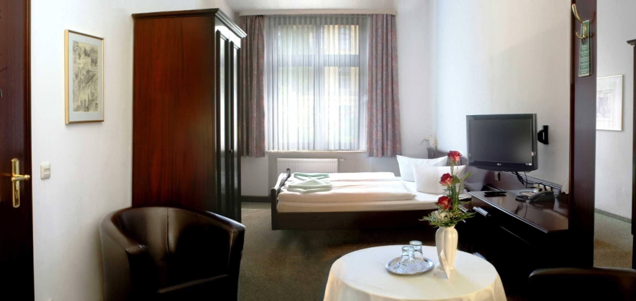 Quarto de hotel com cama, escrivaninha com televisão, cadeira, mesa pequena com vaso e janela com cortinas.