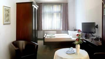 Quarto de hotel com cama, escrivaninha com televisão, cadeira, mesa pequena com vaso e janela com cortinas.
