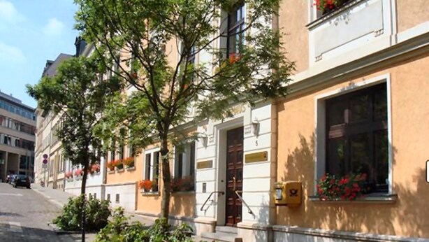 Straßenansicht mit einem Gebäude, das ein großes Fenster, einen Baum und eine kleine Gartenanlage zeigt.