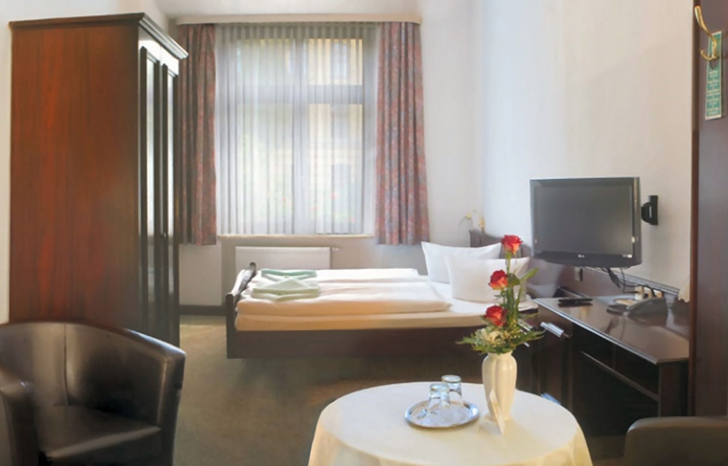 Ein Hotelzimmer mit Bett, Schreibtisch mit Fernseher, Stuhl, Tisch mit Vase und Fenster mit Vorhängen.