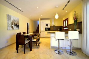 A modern kitchen with a dining area, featuring a wooden dining table, chairs, a bar area with stools, and a staircase leading to an upper level.