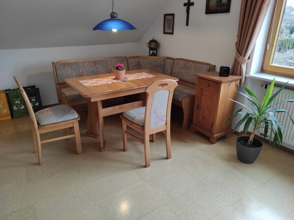 Un comedor con una mesa y sillas de madera, una lámpara colgante azul, una planta en maceta y un radiador.