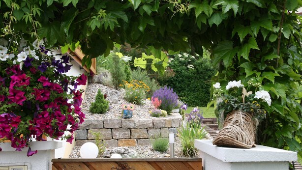 A garden with a variety of flowers and plants, a stone wall, and a wooden fence with a sign. - Apartment on the Rotdornweg (Heiligenstadt i.OFr.)