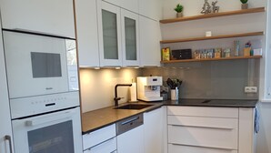 Ett modernt kök med vita skåp, en inbyggd ugn, en kaffemaskin och hyllor i trä med köksredskap.