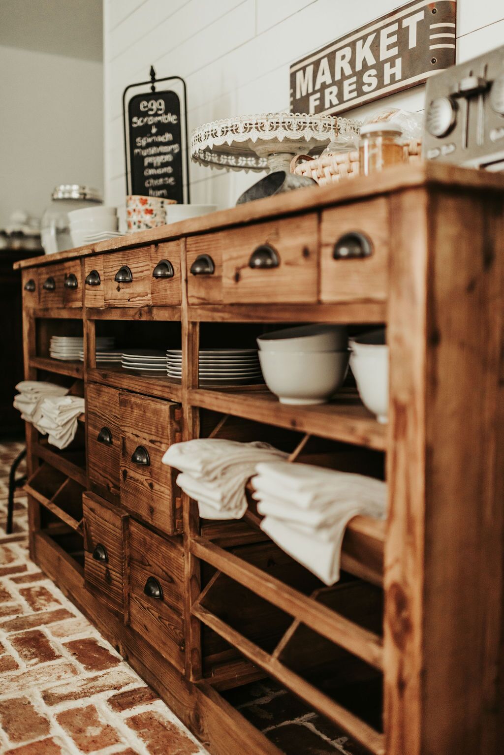 A wooden kitchen counter with multiple shelves and drawers, a sign indicating fresh market produce, and a chalkboard menu listing items like scrambled eggs and spinach.