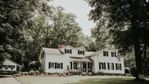 A large, two-story white house with a front porch, surrounded by lush greenery and trees.