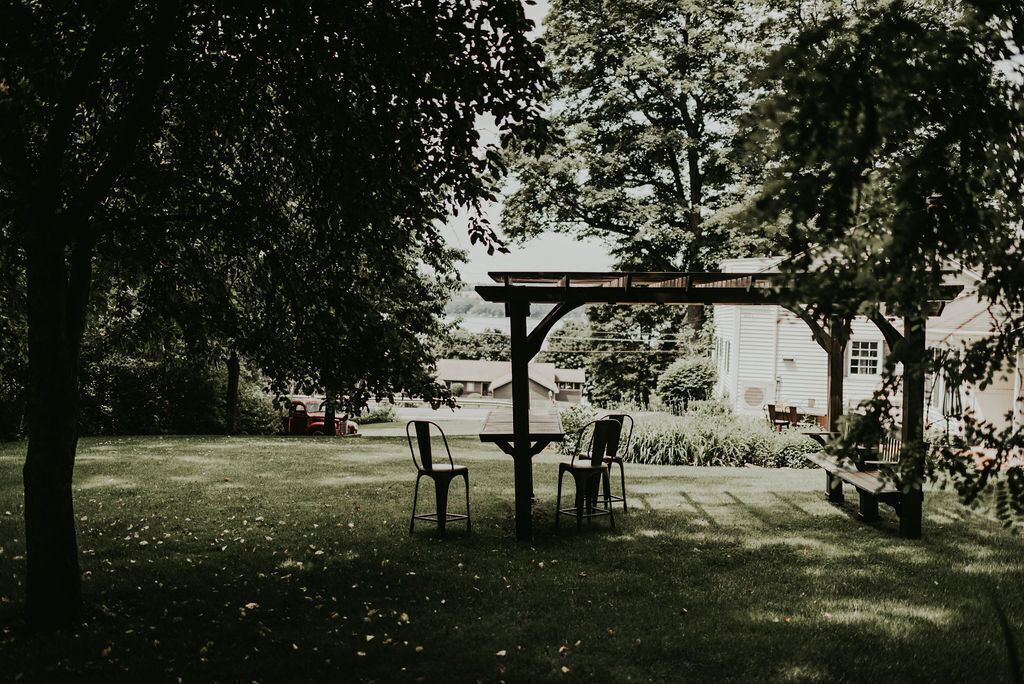 A gazebo with a table and chairs in a grassy area, surrounded by trees and a building in the background.