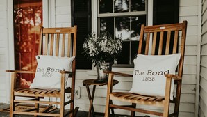 Two rocking chairs with white cushions labeled "The Bond 1835" placed on a porch with a potted plant between them.
