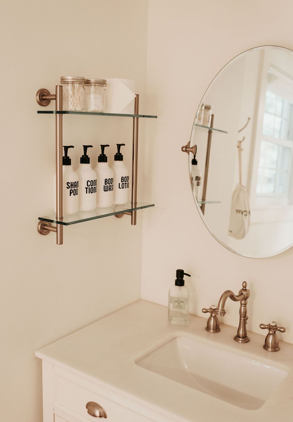 A bathroom with a white vanity, a glass shelf with toiletries, a round mirror, and a brass faucet.